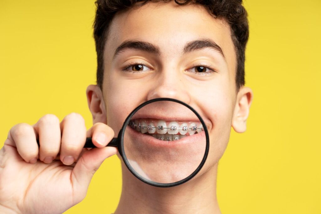 Teen with braces holding a magnifying glass over their smile during early orthodontic evaluation